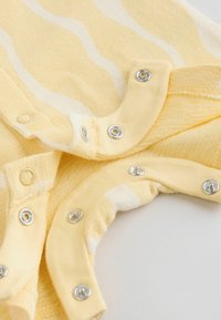 Primo piano di un tessuto a strisce gialle e bianche con bottoni automatici color argento, che mostra il dettaglio della chiusura su materiale morbido e strutturato.