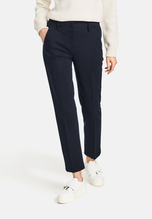 Femme portant un pantalon droit bleu marine, un pull crème et des baskets blanches à bandes velcro, debout avec une main dans la poche.