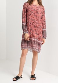 Vestido de manga larga, hasta la rodilla, en color coral con estampados florales, textura plisada y acentos similares a encaje cerca del dobladillo. Combinado con sandalias negras.
