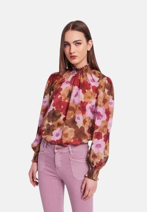 Donna che indossa una camicetta floreale a maniche lunghe con colletto arricciato e pantaloni rosa a vita alta, in piedi su sfondo bianco.