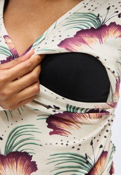 Χέρι που τραβάει προς τα πλάγια το τοπ με σχέδιο λουλουδιών για να αποκαλύψει τη μαύρη εσωτερική στρώση κάτω από την περιοχή του άνω στήθους.