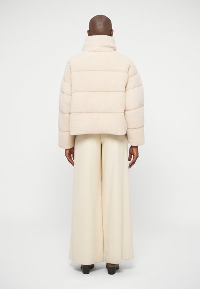 Cream-colored puffer jacket with a high collar, featuring a textured, plush fabric and horizontal quilted design. Worn with wide, cream trousers.