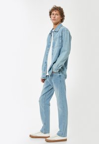 Giacca di jeans azzurro chiaro con bottoni, abbinata a jeans azzurro chiaro. Il modello indossa una maglietta bianca e sneakers, mostrando un look casual e rilassato.