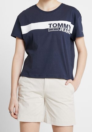 Mørkeblå bomulds T-shirt med en hvid vandret stribe og "TOMMY JEANS" logo. Kombineret med lyse beige shorts, som viser et afslappet look.