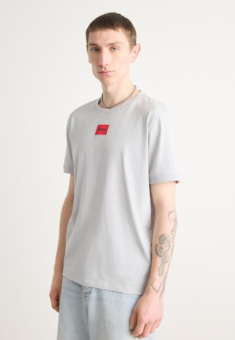 T-shirt in cotone grigio chiaro con scollo rotondo e maniche corte, caratterizzato da un logo rosso a forma di quadrato ben visibile sul petto. Vestibilità casual.
