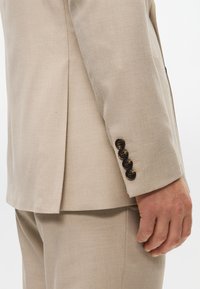 Beige Sakko mit taillierter Passform, glatter Textur, vier dunklen Knöpfen am Ärmel und einem Schlitzdetail am Rücken.