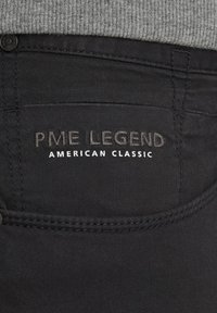 Jeans de denim negro con un acabado texturizado, que cuentan con una cinturilla de tela gris. El bolsillo trasero muestra "PME LEGEND" y "AMERICAN CLASSIC" en costura.