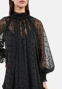 Schwarze durchsichtige Bluse mit Punkten, hohem gerafftem Kragen und voluminösen Ärmeln. Mit rüschiertem Saum und strukturierter Stoffqualität.