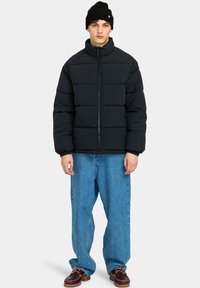 Giacca a vento nera con colletto alto e chiusura con zip, abbinata a jeans blu a vestibilità ampia e scarpe marroni con lacci. Accessorio berretto nero.
