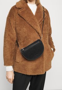 Borsa a tracolla in suede nero con hardware in argento, forma semicircolare e dettagli testurizzati. Indossata sopra un cappotto marrone testurizzato.