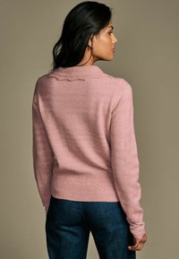 Medvilnės mišinys rožinis megztinis su raukšlėtu apačiu ir bangomis primenančiu iškirpimu. Ilgos rankovės su sudėtingais detalėmis ant rankogalių.