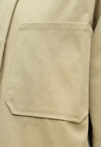 Khaki-skjorte med en stor, firkantet lomme på venstre side, lavet af slidstærkt stof med en glat tekstur og synlige syningsdetaljer.