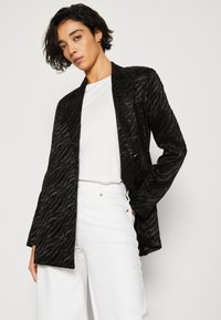 Svart texturerad blazer med zebramönster, öppen front och långa ärmar, i kombination med en vit t-shirt och vida vita byxor.