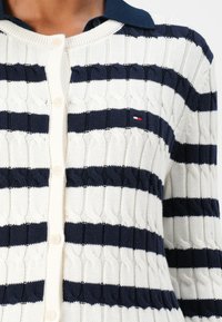 Marineblå og kremstripet strikket cardigan med kabelmønster, knappelukking og marineblå krage. Har liten logo på venstre bryst.