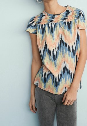 Frau trägt eine kurzärmelige Bluse mit Zickzack-Muster in Pfirsich, Blau, Schwarz und Creme sowie eine dunkelgraue Jeans vor einer hellblauen Wand.