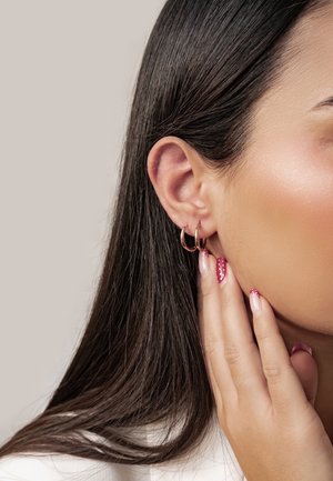 Kobieta o długich ciemnych włosach, nosząca dwie małe złote kolczyki w kształcie koła i dotykająca twarzy zadbanymi paznokciami w kolorze różowym z białymi kropkami.