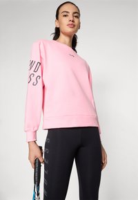 Sweatshirt rose clair avec col rond et lettrage noir sur la manche, associé à un legging noir avec du texte gris sur le côté.