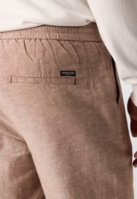 Pantalons en lin marron avec une taille élastique, dotés d'une poche arrière et d'une petite étiquette noire. La texture est légèrement texturée et légère.