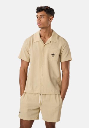 Homme portant une chemise beige texturée à manches courtes boutonnée et un short assorti avec de petits logos noirs de palmiers, mains dans les poches, regardant vers la gauche.