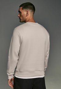 Sudadera beige clara hecha de una tela suave, con un cuello redondo, puños de canalé y un dobladillo recto, mostrada desde la parte de atrás.