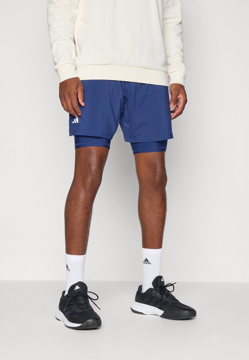 Homme portant un sweat-shirt blanc, un short bleu superposé, des chaussettes blanches Adidas et des baskets noires Adidas, debout sur un sol blanc.