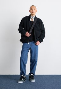 Giacca in pile nera con zip sopra una maglia nera, jeans blu larghi e sneakers nere/grigie. Gli accessori includono un bandana bianca legata intorno al collo.