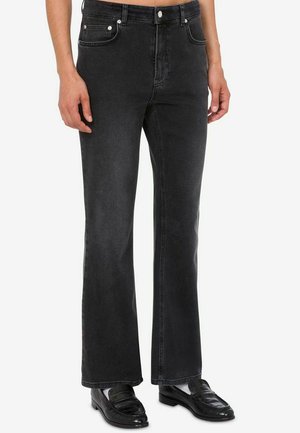Uomo che indossa jeans bootcut neri e mocassini in pelle nera con calzini bianchi, in piedi davanti a uno sfondo bianco uniforme.