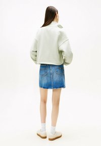Sweatshirt cropped di un verde chiaro con un colletto alto, abbinato a una minigonna di denim blu e sneakers bianche, vista da dietro.
