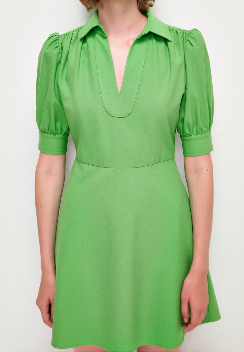 Robe verte avec un col à revers, des manches courtes bouffantes et une taille cintrée qui s'évase. Confectionnée en tissu lisse uni.