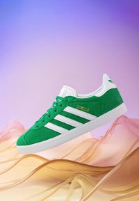 Green suede sneaker with white leather accents and three striped detail. Rounded toe, flat white rubber sole, and gold branding "GAZELLE."