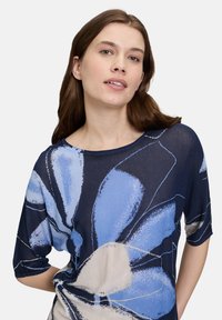 Niebieska bluzka w kwiatowy nadruk z krótkimi rękawami, wykonana z lekkiego materiału, okrągłym dekoltem oraz abstrakcyjnymi wzorami kwiatowymi w odcieniach niebieskiego i kremowego.