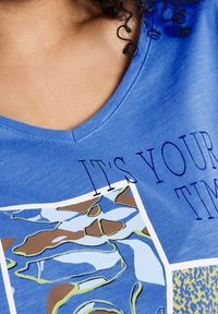 T-shirt in cotone blu con scollo a V, stampa grafica astratta in marrone e verde, e la scritta "È IL TUO MOMENTO" in blu scuro.