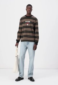 Camisa de manga larga a rayas en negro, beige y rosa con un logo central, combinada con unos jeans de mezclilla azul claro y zapatos negros, sosteniendo una bolsa tipo tote.