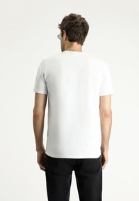 Camiseta blanca de manga corta con cuello redondo, textura suave y un diseño simple, combinada con jeans oscuros. La vista muestra la parte posterior.
