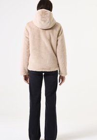 Chaqueta de forro polar beige con capucha, con una textura suave y un ajuste holgado, combinada con pantalones de mezclilla oscuros. La vista es desde la parte posterior.