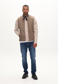 Beige quiltad väst över en ljusbrun tröja, kombinerad med blå jeans och svarta skor. Accenterad med en svart bandana runt halsen.