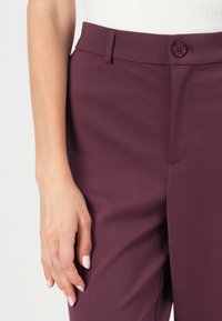 Burgundské kalhoty z hladkého materiálu, s tlačítkovým zapínáním, přiléhavým střihem a plochým předním designem. Ruka položená na boku.