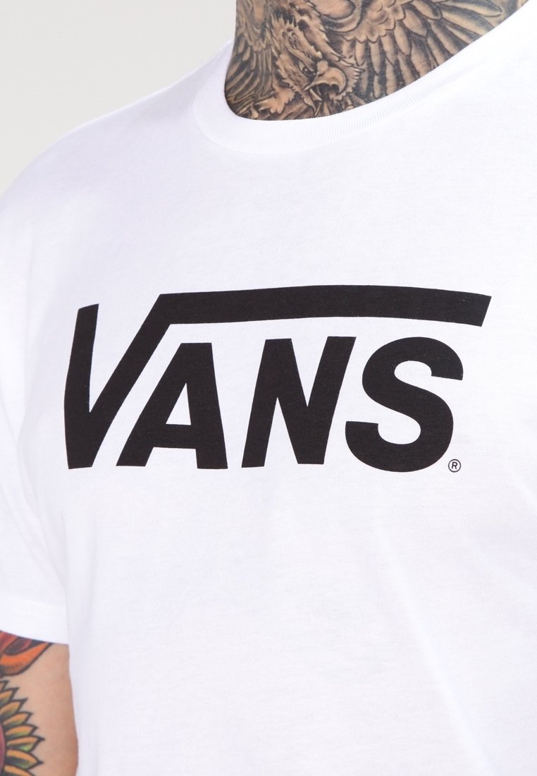 T-shirt blanc avec un grand logo noir Vans sur la poitrine, porté par une personne avec des tatouages détaillés au cou et aux bras.