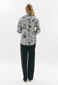 Eine Frau mit langen blonden Haaren steht mit dem Rücken zur Kamera und trägt eine langärmlige gestreifte Bluse mit Blumendruck und schwarze weitgeschnittene Hosen vor einem weißen Hintergrund.
