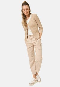 Beige Rippstrick-Cardigan mit Knöpfen, weißem T-Shirt und beigen Cargo-Hosen. Die Sneakers bestehen aus einer Mischung aus beigen und weißen Materialien. Lässiges Outfit.