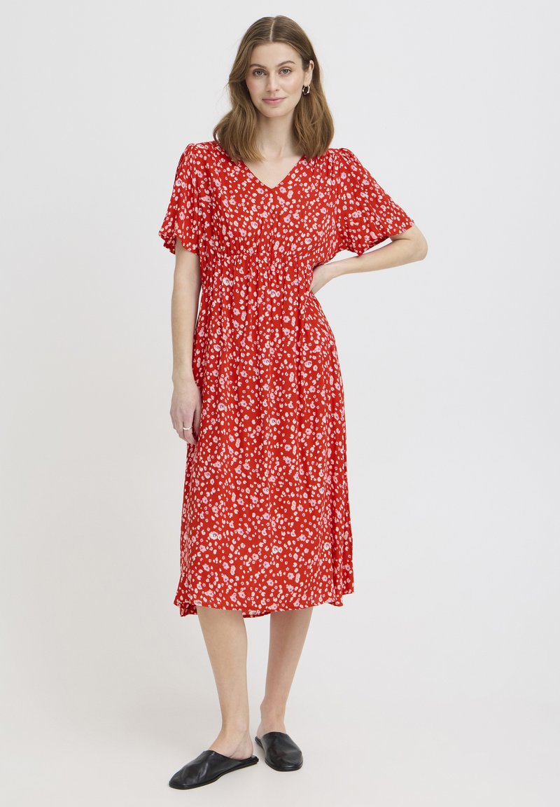 Femme portant une robe midi rouge à motif floral blanc et manches courtes, debout avec une main sur la hanche et des chaussures noires sans lacets.