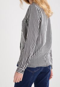 Camisa de manga larga con rayas blancas y negras, bolsillo frontal, puños con botones y corte holgado, combinada con vaqueros azules.