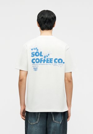 Person med kort mørkt hår iført hvid t-shirt med blå teksten "SOL COFFEE CO." og blomsterprint på ryggen, kombineret med mørke denimjeans.