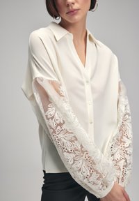 Cream blouse with a collar, button front, and transparent lace sleeves featuring floral embroidery and delicate detailing. Black pants are also visible.