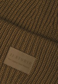 Tissu en tricot côtelé marron avec une étiquette en cuir rectangulaire cousue portant l'inscription "JJ STUDIO Our Legacy Your Future."