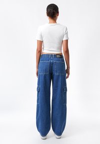 Pantalones cargo de mezclilla azul claro con piernas anchas, dos bolsillos laterales y una etiqueta negra en la parte posterior. Combinados con un top corto blanco ajustado.