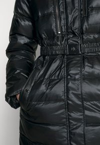 Svart puffjacka med en glänsande yta, åtsittande midja, fickor med dragkedja och ett bälte med metallspänne. Har en quiltad textur.