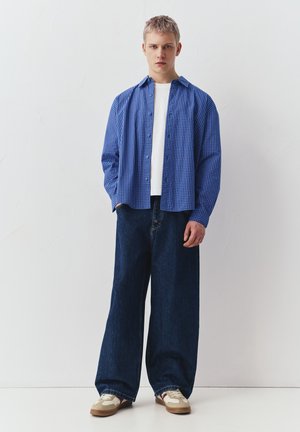 Mladić stoji s rukama u džepovima, nosi plavu košulju na kockice, bijelu majicu, široke tamne traperice i bež tenisice.