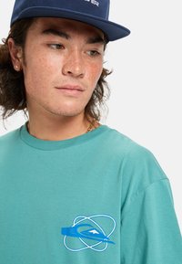 Joven con cara pecosa que lleva una gorra azul y una camiseta verde azulado con un logo abstracto azul en el pecho, mirando hacia un lado.