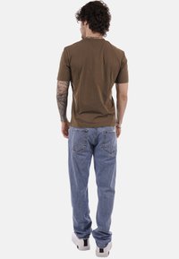 Uomo con capelli ricci, indossa una maglietta marrone a maniche corte, jeans blu e scarpe da ginnastica bianche, in piedi di spalle su sfondo bianco.